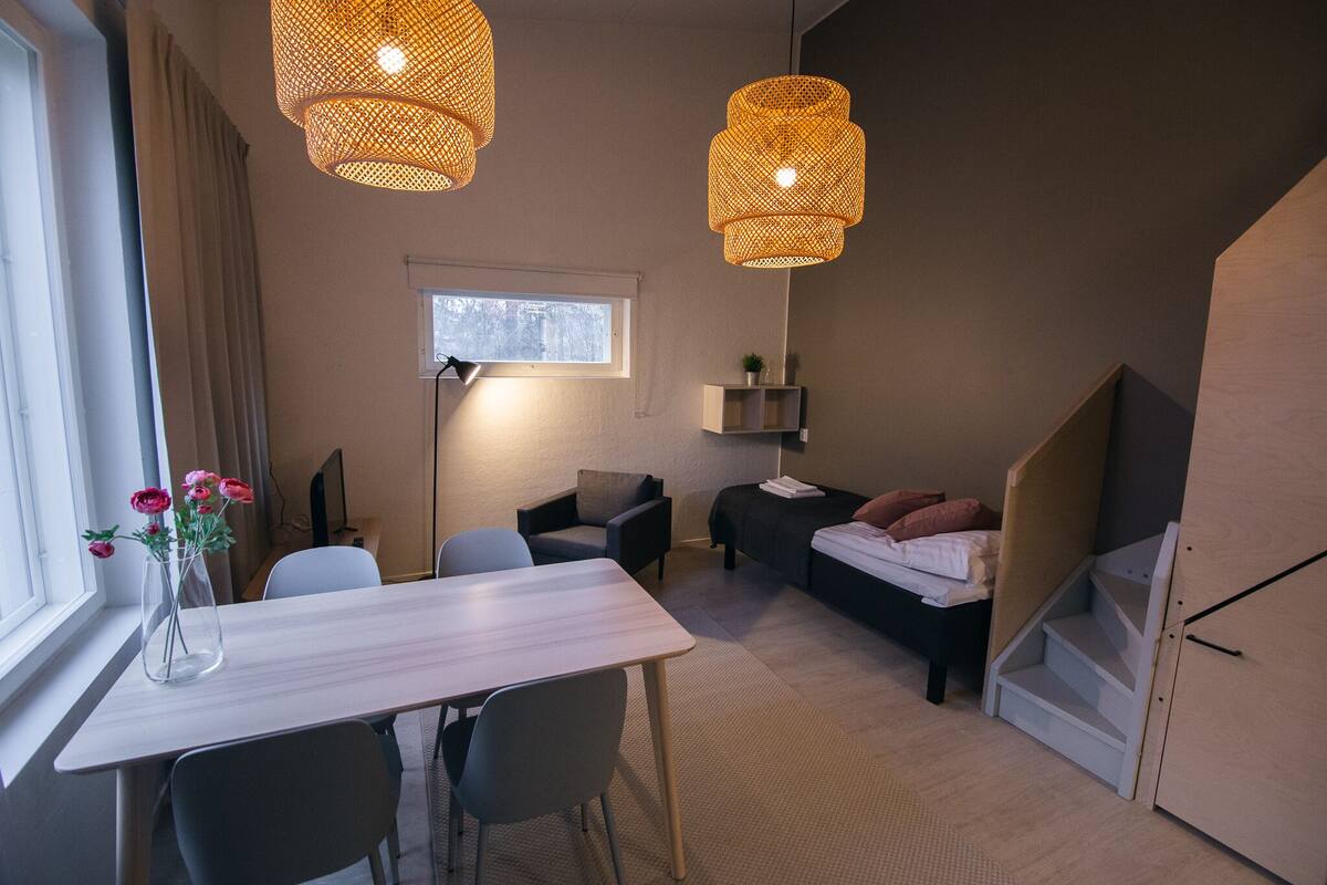 A modern room with a dining table, chairs, a sofa, and a bed. There are two hanging lights and a window with curtains.