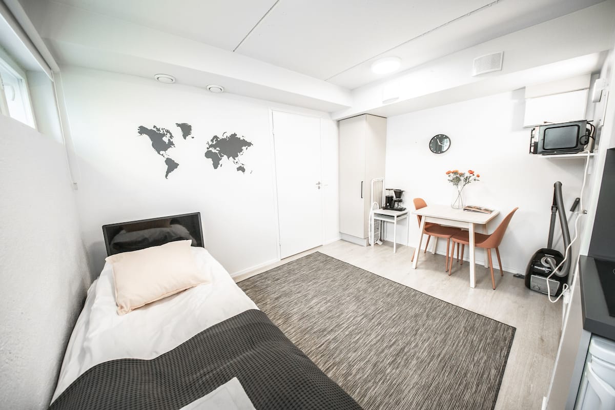 A modern hotel room with a bed, a desk, a chair, a small table, a world map on the wall, a clock, and a microwave.
