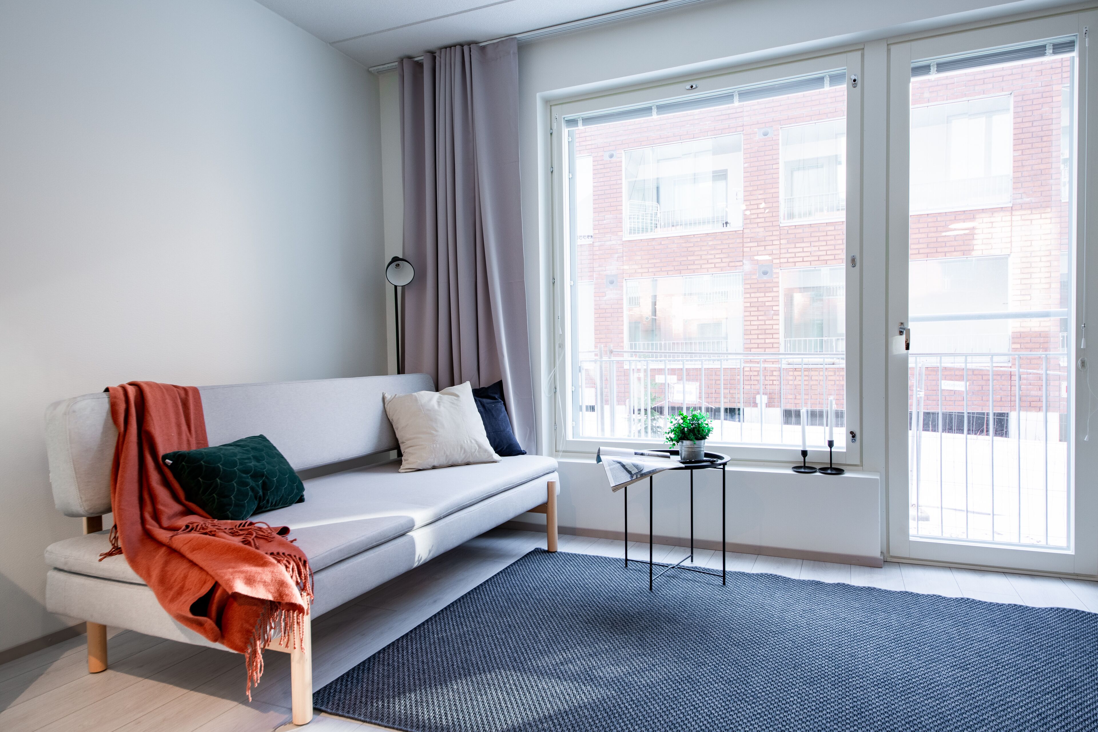 A modern living room with a sofa, a small side table, a potted plant, and a view of a brick building through large windows.