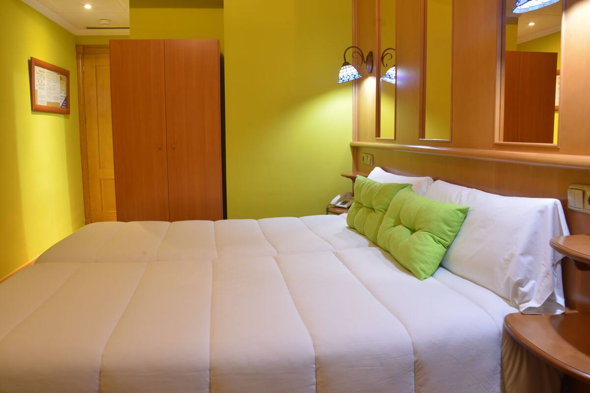 A neatly made bed with white bedding and a green pillow, a wooden wardrobe, and a mirror above the bed.