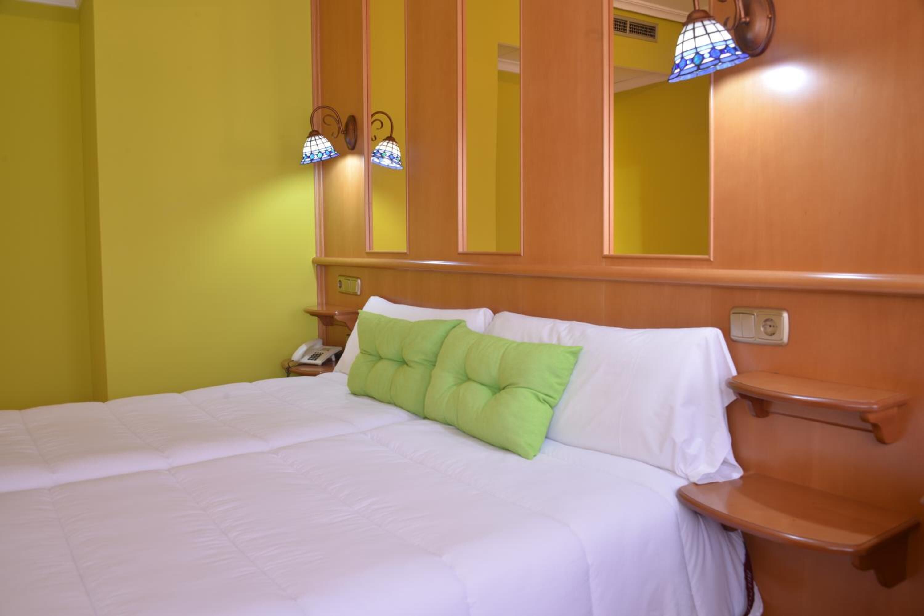 Habitación de hotel con cama, almohadas verdes, cabecero de madera, mesitas de noche y lámparas de pared.