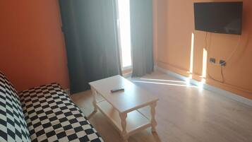Una habitación con un sofá a cuadros, una mesita blanca, un televisor en la pared y una ventana con cortinas.