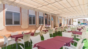 An outdoor dining area with tables and chairs, a covered walkway, and a building with windows. - Hotel Aros (Casas-Ibanez)