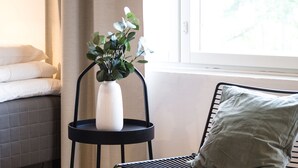 A room with a bed, a small black metal chair, a white vase with a plant, and a window with curtains.