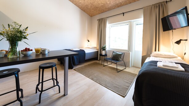 A modern room with a bed, a desk with stools, a small table with a vase of flowers, and a TV mounted on the wall.