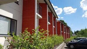 A red building with multiple windows and balconies, a black car parked in front, and green shrubs in the foreground.