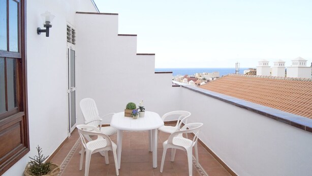 Ein Balkon mit einem weißen Tisch und Stühlen, einem Korbwannenstuhl mit einer Pflanze und Blick auf das Meer und Gebäude in der Ferne.
