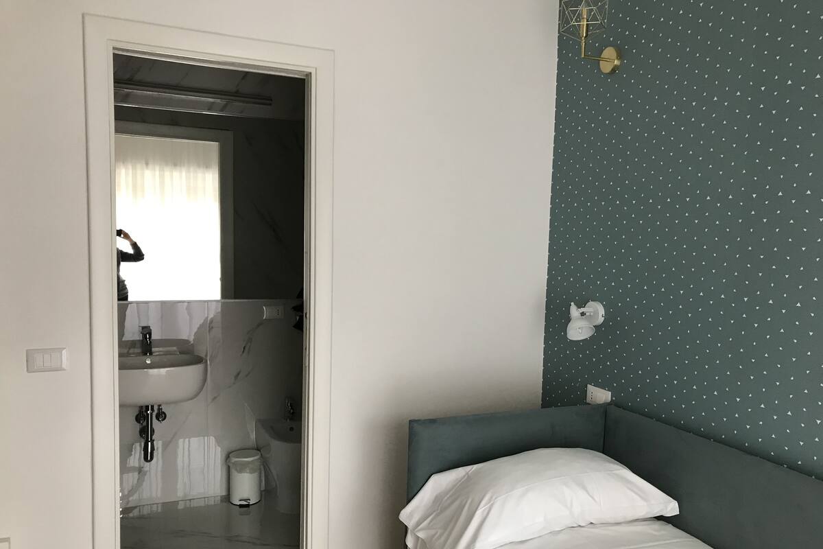 Une chambre à coucher avec un lit, une table de chevet, une lampe fixée au mur et une salle de bain visible à travers une porte ouverte.