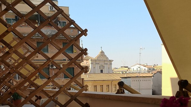 A terrace with a wooden lattice, potted plants, a white table, and a view of buildings in the background. - Affreschi Al Vaticano (Rome)