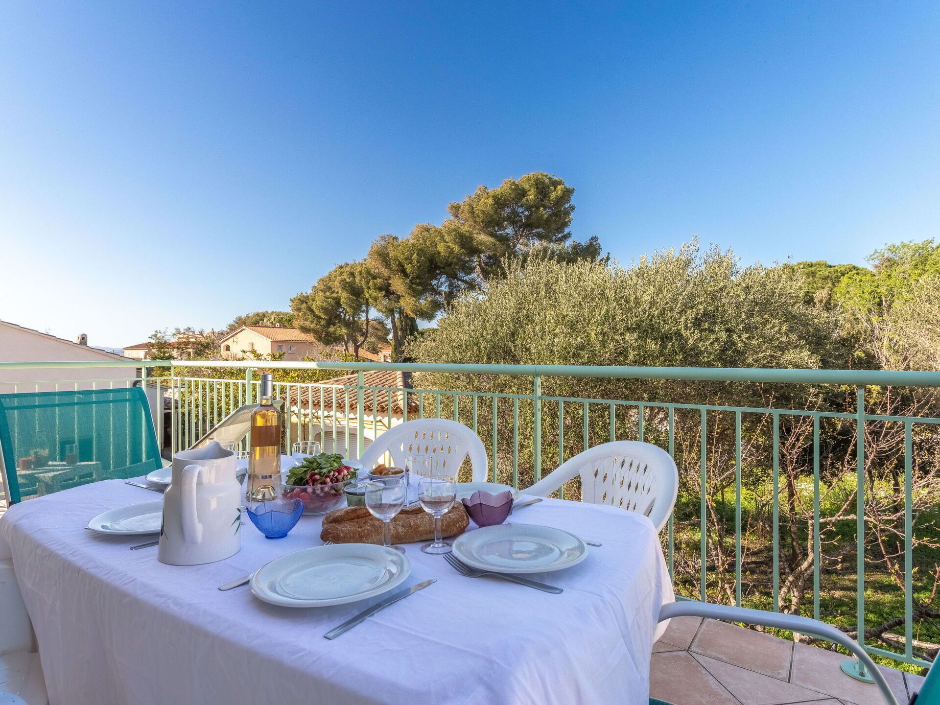 A table set outdoors with a white tablecloth, plates, cutlery, a bottle, glasses, and a loaf of bread.