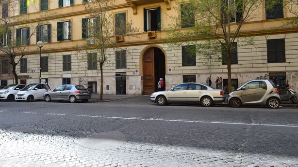 Ein mehrstöckiges Gebäude mit einem zentralen Eingang, Autos, die an der Straße parken, und Bäume davor.
