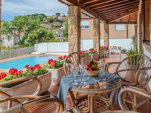 A covered outdoor dining area with wicker chairs, a pool, and red flowers.