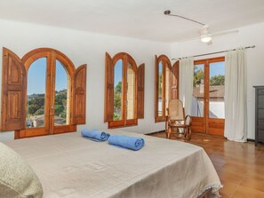 A room with a bed, a chair, wooden-framed windows, a ceiling fan, and a view of the outside.