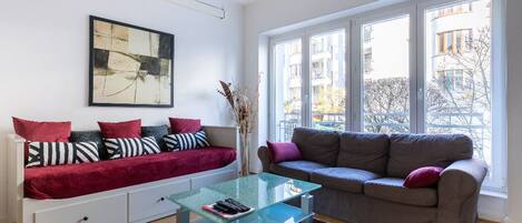 A modern living room with a glass coffee table, a sofa with patterned cushions, and a large window offering a view of buildings and trees.