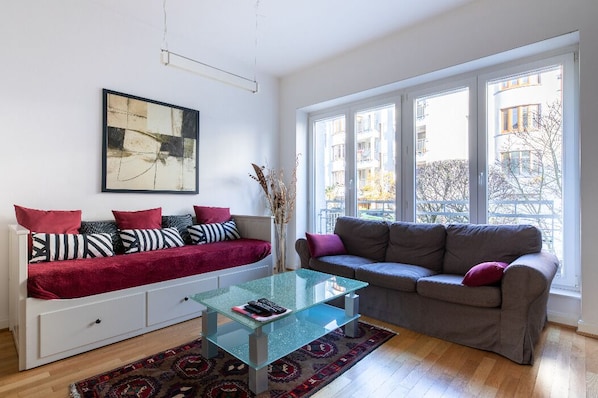 A modern living room with a glass coffee table, a sofa with patterned cushions, and a large window offering a view of buildings and trees.