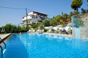 A swimming pool with lounge chairs and umbrellas, surrounded by a stone wall and a building with balconies. - Filia Studios & Apartments (Skiathos)