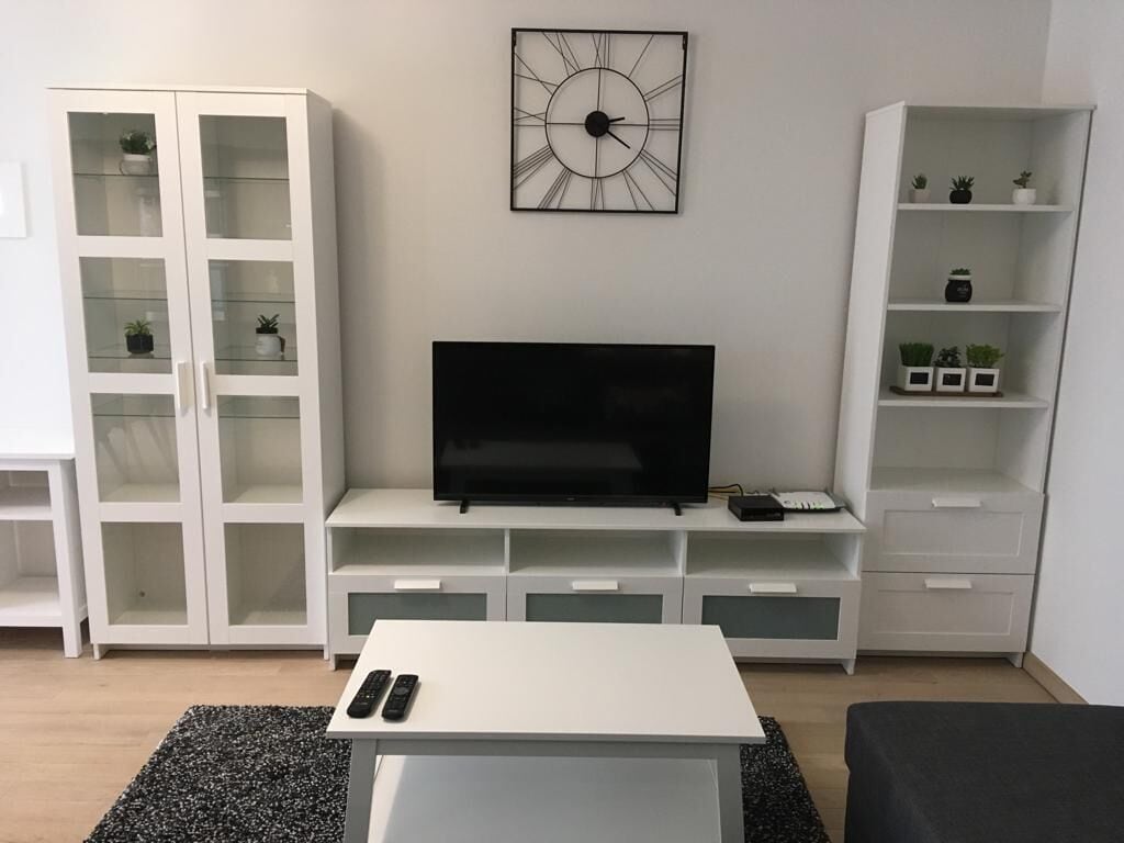 Un soggiorno con una TV a schermo piatto su un mobile bianco, un armadietto bianco con ante in vetro e un orologio appeso al muro.