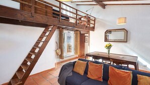 A loft-style room with a wooden staircase, a sofa with cushions, a dining table with chairs, and a mirror on the wall.