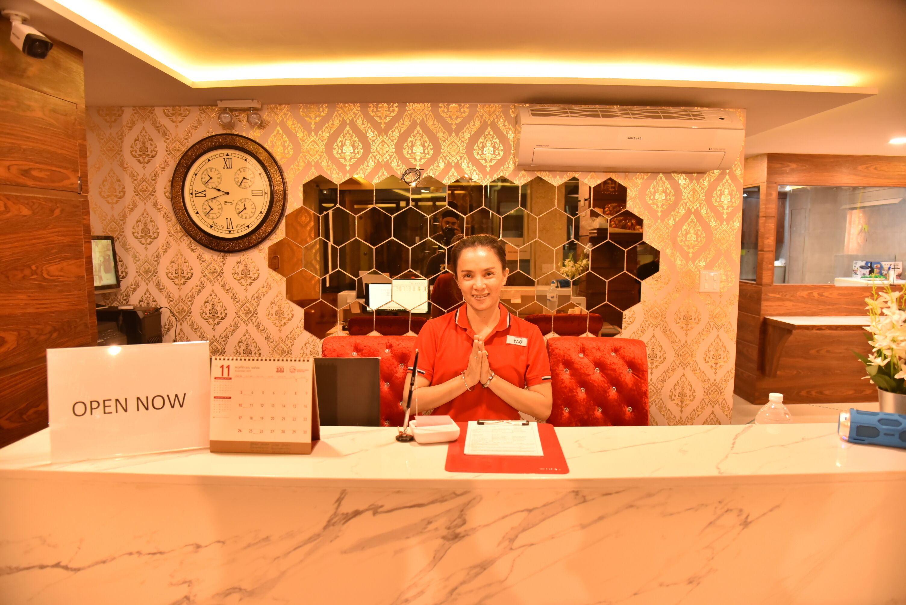 A hotel reception desk with a 'OPEN NOW' sign, a wall clock, and a calendar.