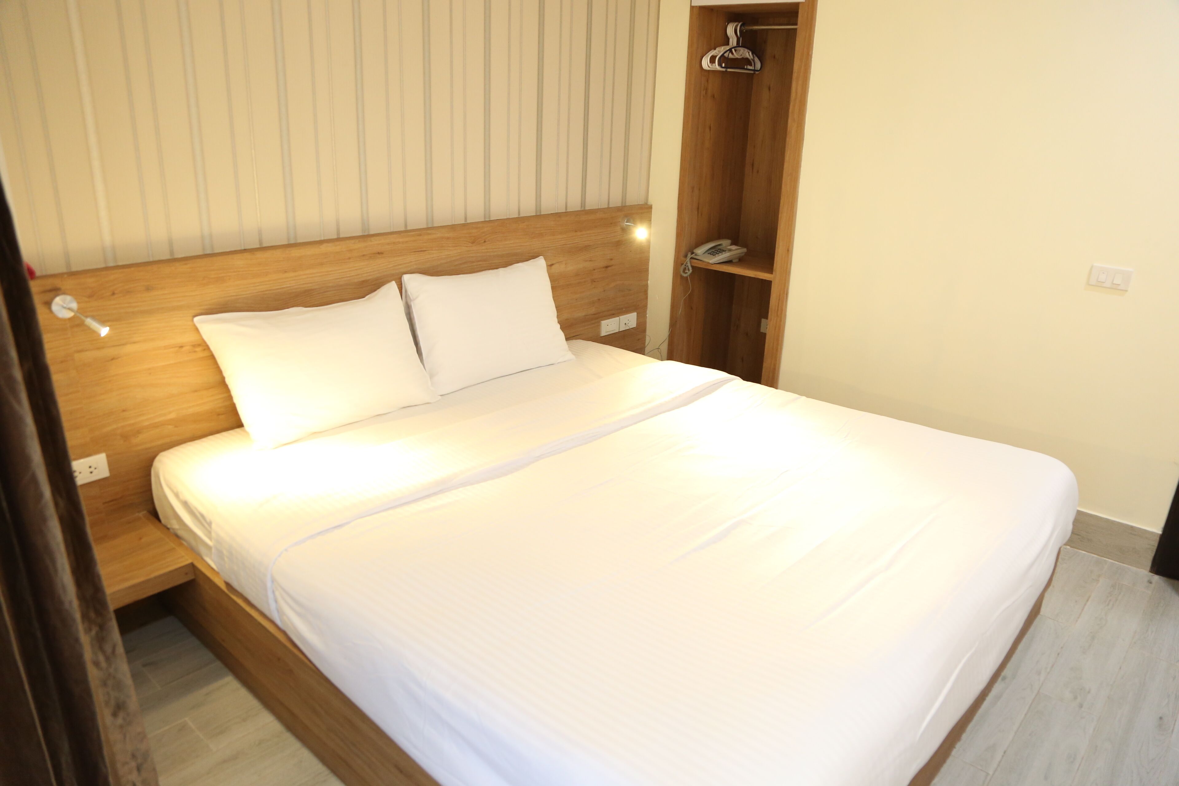 Una cama bien hecha con sábanas blancas y dos almohadas en una habitación con cabecera de madera y un armario de madera con perchero.