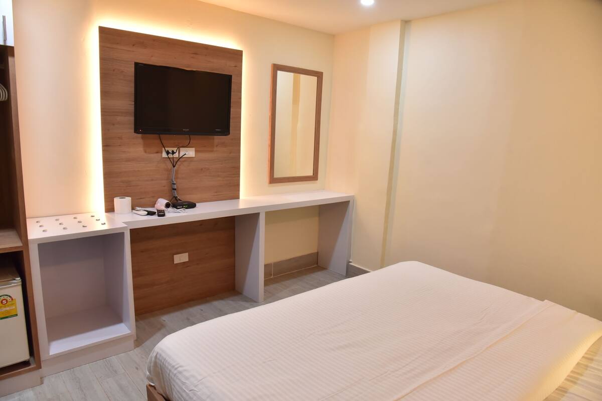 A bedroom with a bed, a desk with a flat-screen TV, a mirror, and a shelf with toiletries.