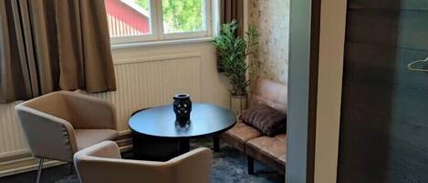 A room with a sofa, two armchairs, a small round table with a vase, a window with curtains, and a view of greenery outside.