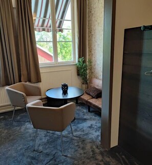 Una habitación con sofá, dos sillones, una mesita redonda con un jarrón, una ventana con cortinas y vistas al exterior a zonas verdes.