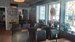 A restaurant interior with wooden tables and black chairs, large windows with potted plants, and a clock on the wall. - Sjugare Bed & Breakfast (Leksand)