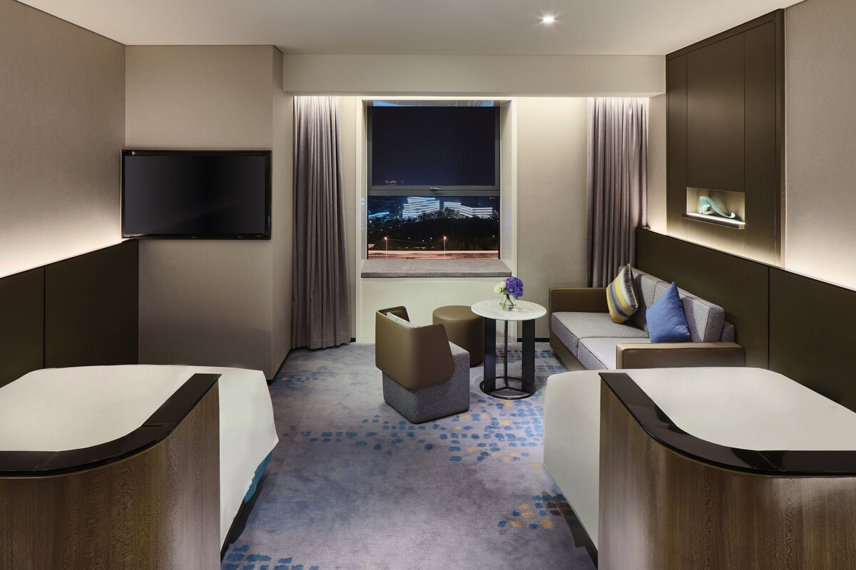 A modern hotel room with a large bed, a sofa, a small table with a vase of flowers, and a TV mounted on the wall.