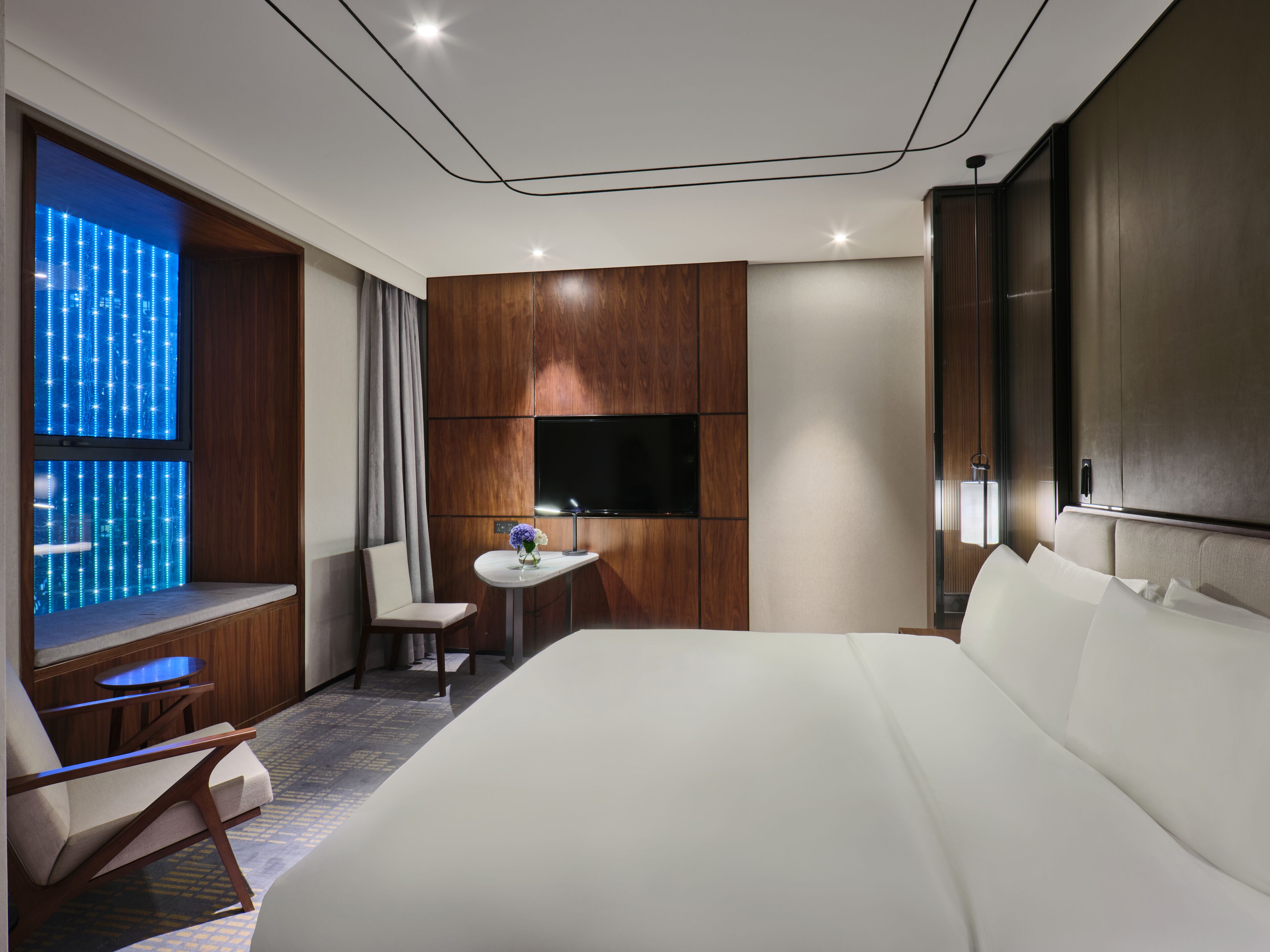 Ein modernes Hotelzimmer mit einem großen Bett, einem an der Wand montierten Fernseher, einem kleinen Tisch mit Blumenvase und einem Fenster mit Blick auf die Stadtlichter.