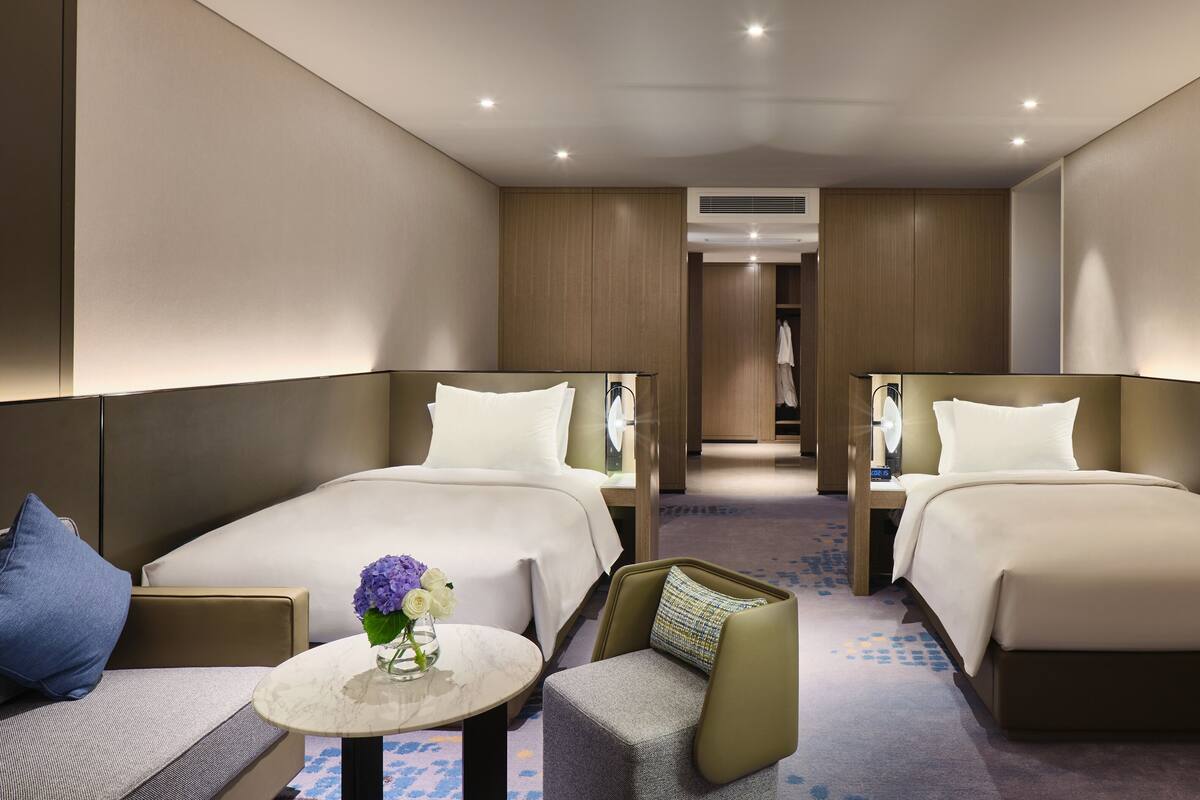 A hotel room with two beds, a small table with a vase, and a chair.