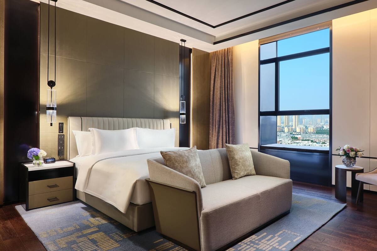 A modern hotel room with a large bed, a sofa, a small table with a vase, and a view of the city through large windows.