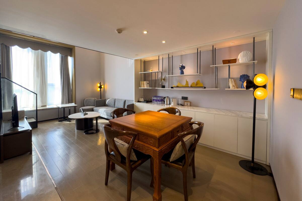 A modern living room with a dining table, chairs, a sofa, and a kitchen area with shelves and a coffee machine.