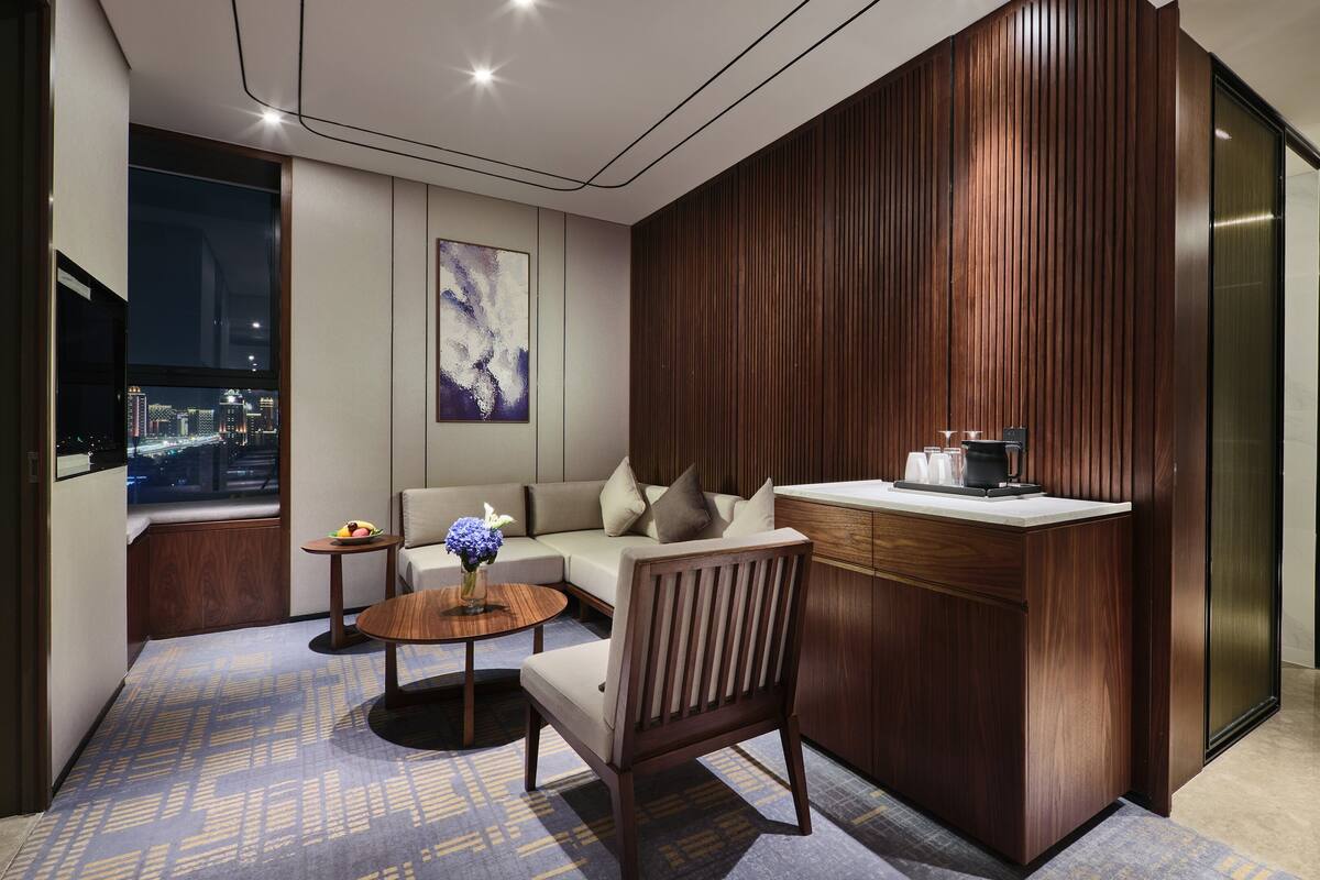 A modern hotel room with a sofa, a small table, a chair, a coffee station, and a city view.