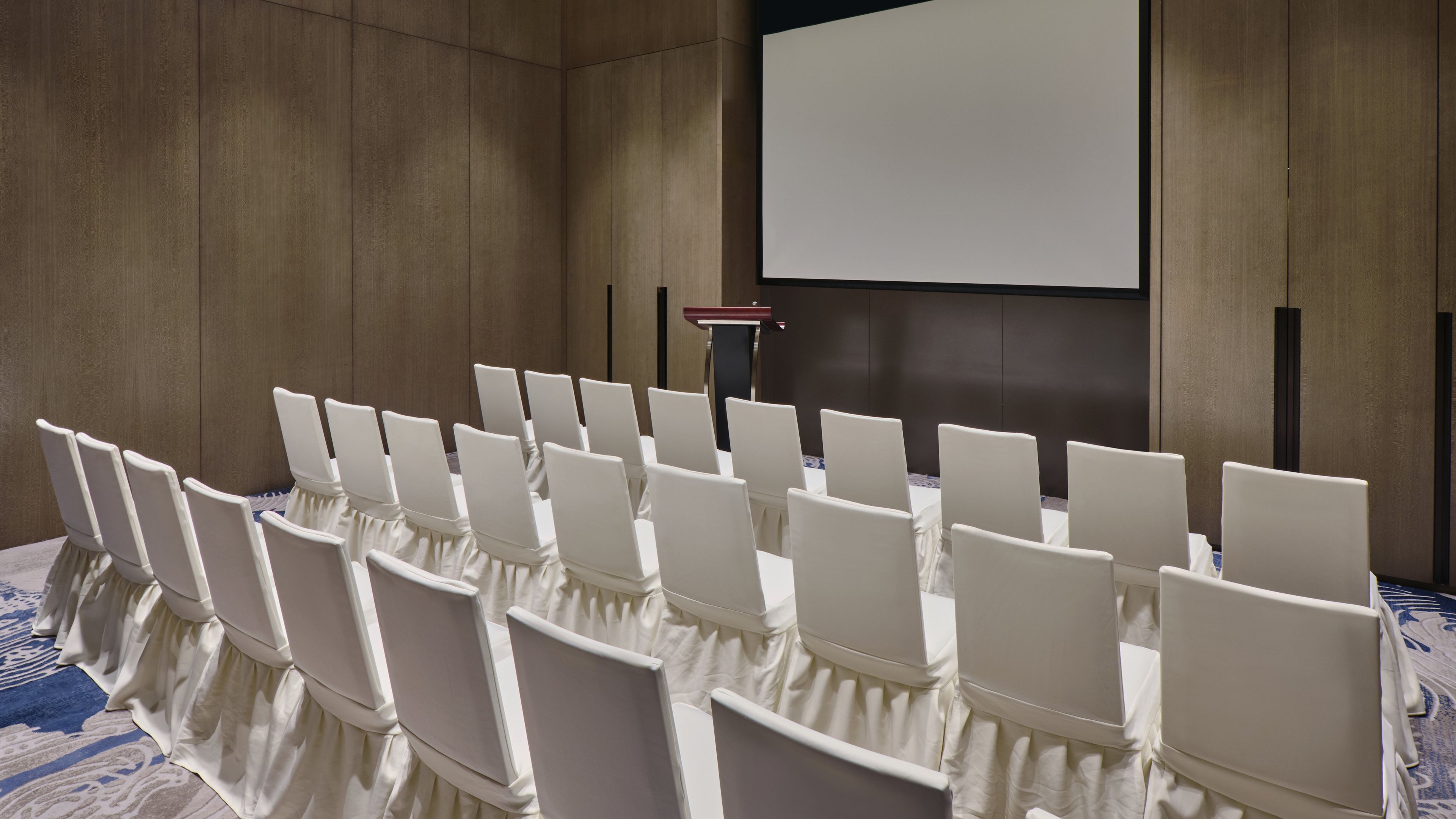 A conference room with rows of white chairs, a podium, and a projection screen.