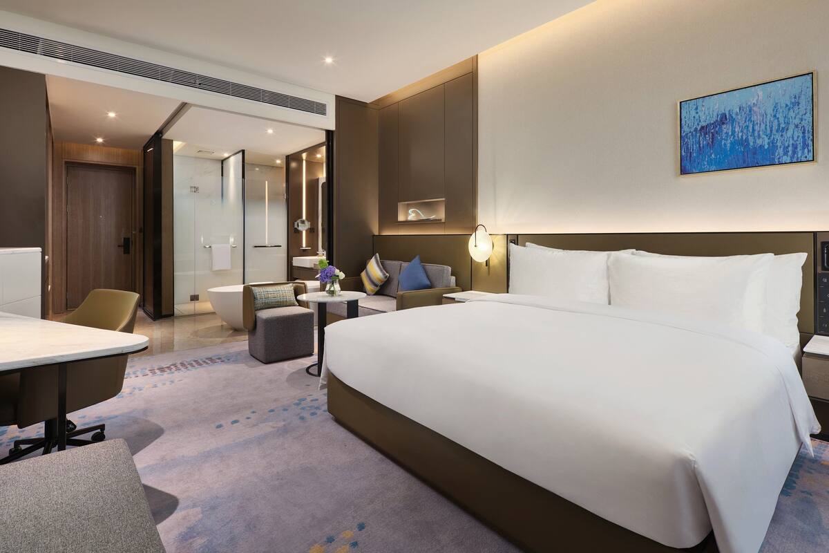 A modern hotel room with a large bed, a desk, a sofa, and a bathroom visible through an open door.