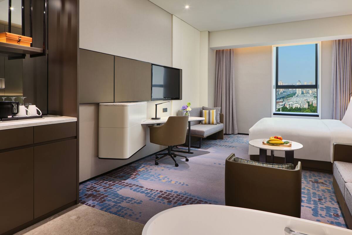 A hotel room with a bed, a desk, a chair, a small table with fruit, a TV, and a view of a cityscape through a window.