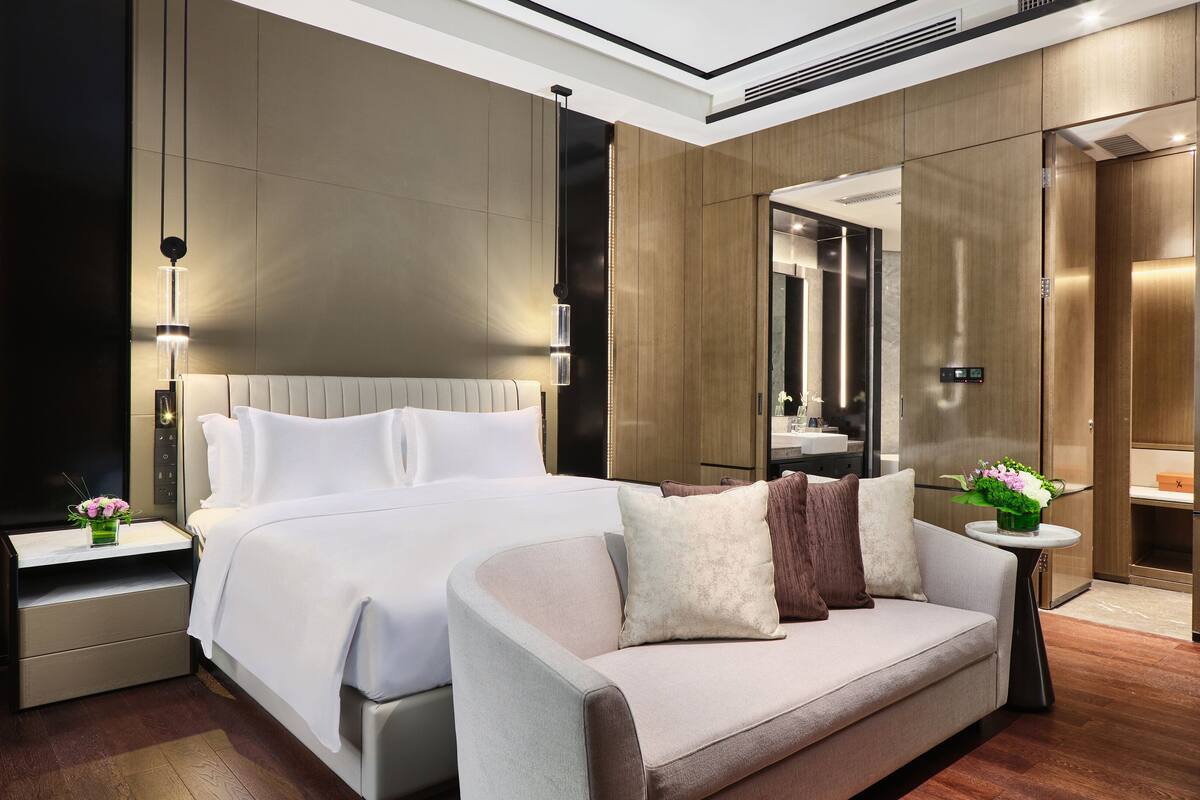 A modern hotel room with a large bed, a sofa, a nightstand, a small table with a vase, and a mirror.