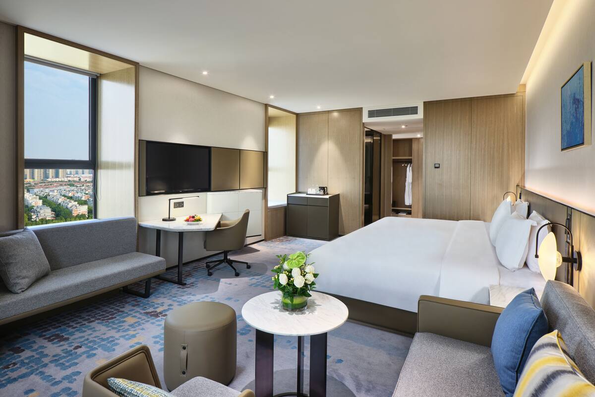 A modern hotel room with a large bed, a sofa, a desk, and a view of the city.