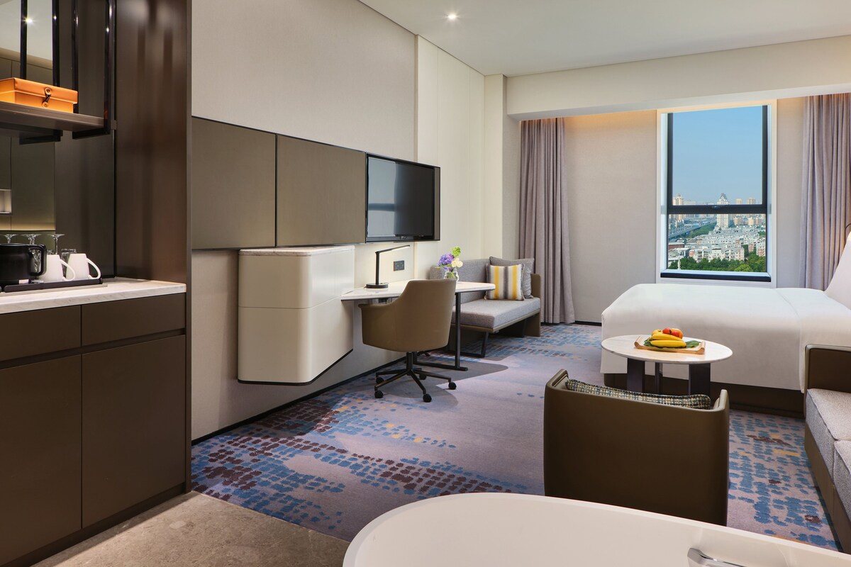 A hotel room with a bed, a desk, a sofa, a small table with fruit, a TV, and a window with a city view.
