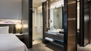 A modern hotel room with a bed, a glass-enclosed shower area, a large mirror, and a bedside table with a lamp. - Crowne Plaza Wuhan Development Zone by IHG (Wuhan)