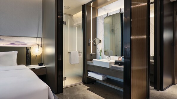 A modern hotel room with a bed, a glass-enclosed shower area, a large mirror, and a bedside table with a lamp. - Crowne Plaza Wuhan Development Zone by IHG (Wuhan)