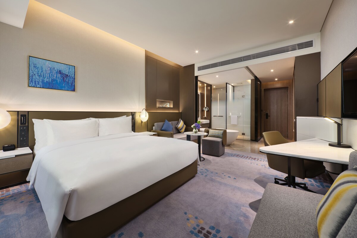 A modern hotel room with a large bed, a desk, a chair, and a bathroom.