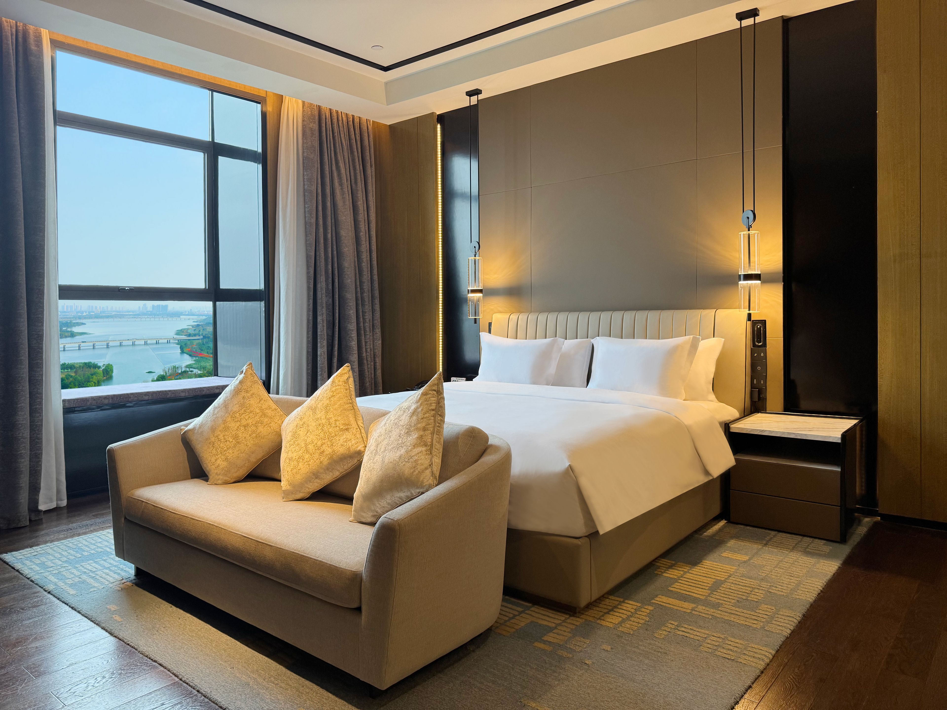A hotel room with a large bed, a sofa, a nightstand, and a view of a cityscape through the window.