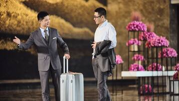 Two men in business attire, one holding a suitcase, in a room with a floral arrangement and a patterned wall.