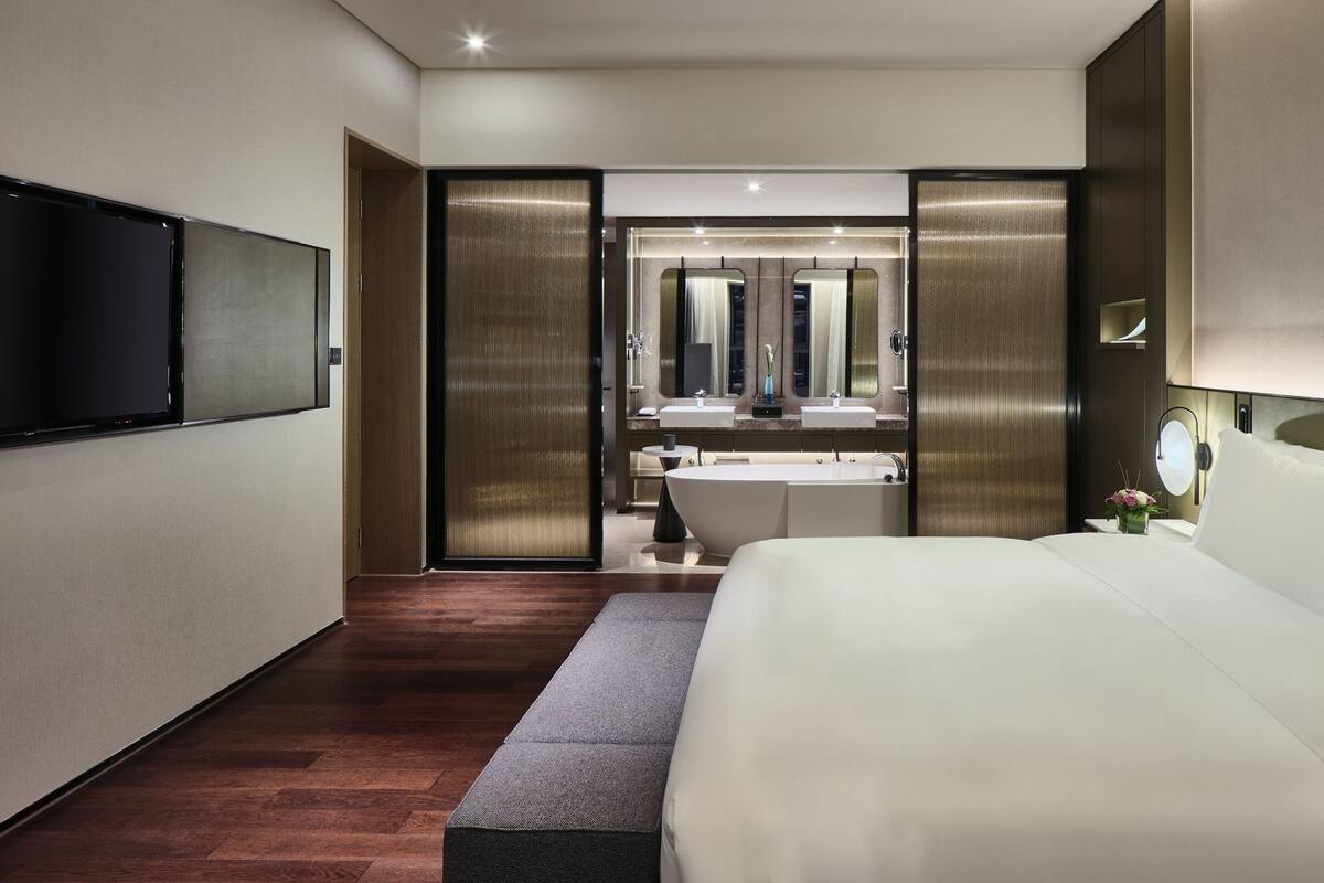 A modern hotel room with a large bed, a flat-screen TV mounted on the wall, a large mirror, and a separate bathroom with a bathtub and sink.