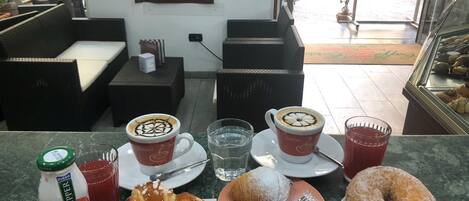 Une scène de café avec deux tasses de café, une bouteille d’eau, un verre de jus de fruits et des pâtisseries sur le comptoir.