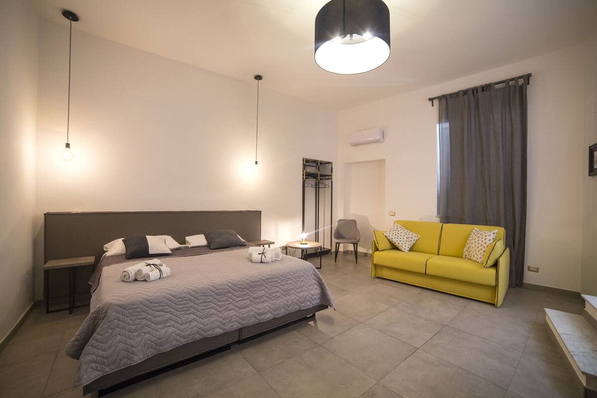 A modern hotel room with a large bed, a yellow sofa, a small table, and a bench.