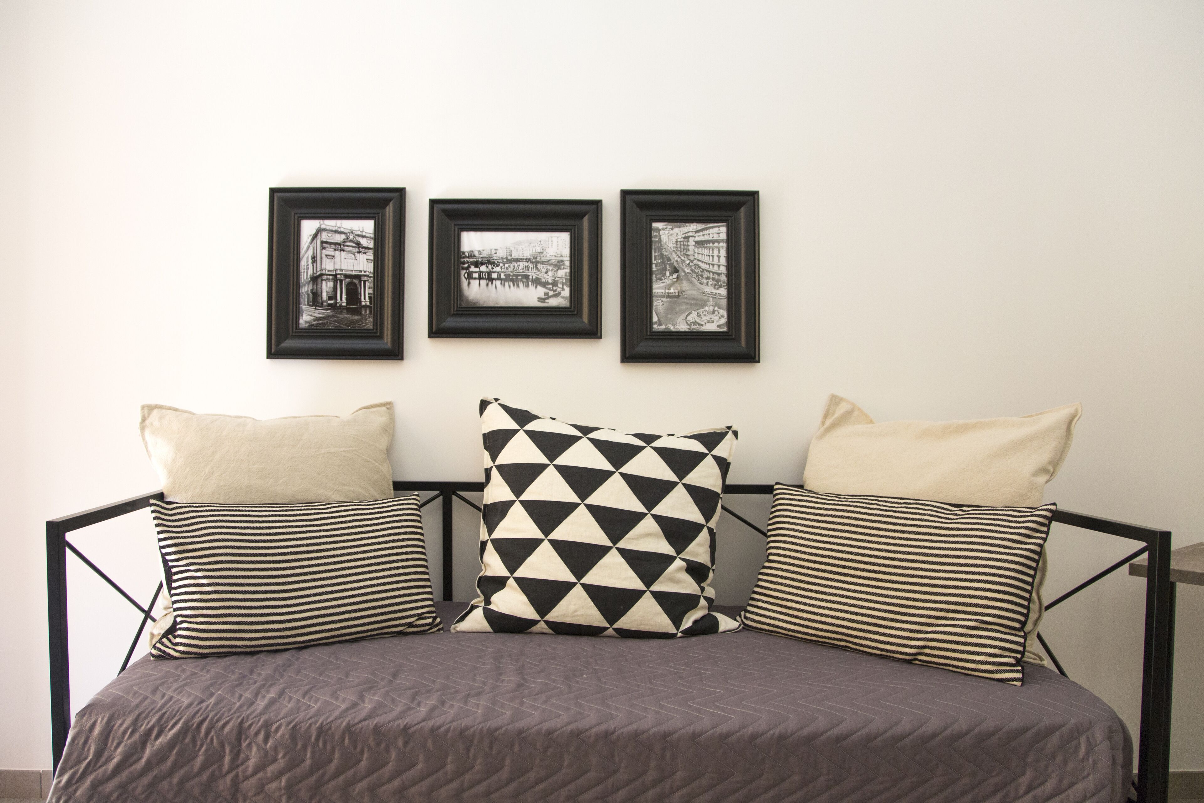 A bed with a gray quilt, three pillows (one striped, one with a black and white checkered pattern, and one plain), and three framed black and white photographs on the wall above.