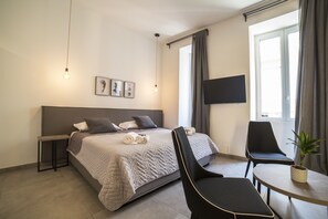 Een moderne hotelkamer met een bed, twee stoelen, een tafeltje, een televisie en een raam met gordijnen.
